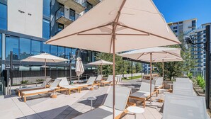 A rooftop lounge area with sun loungers and umbrellas, a pool, and modern buildings in the background. - Saiya  804 (Phoenix)