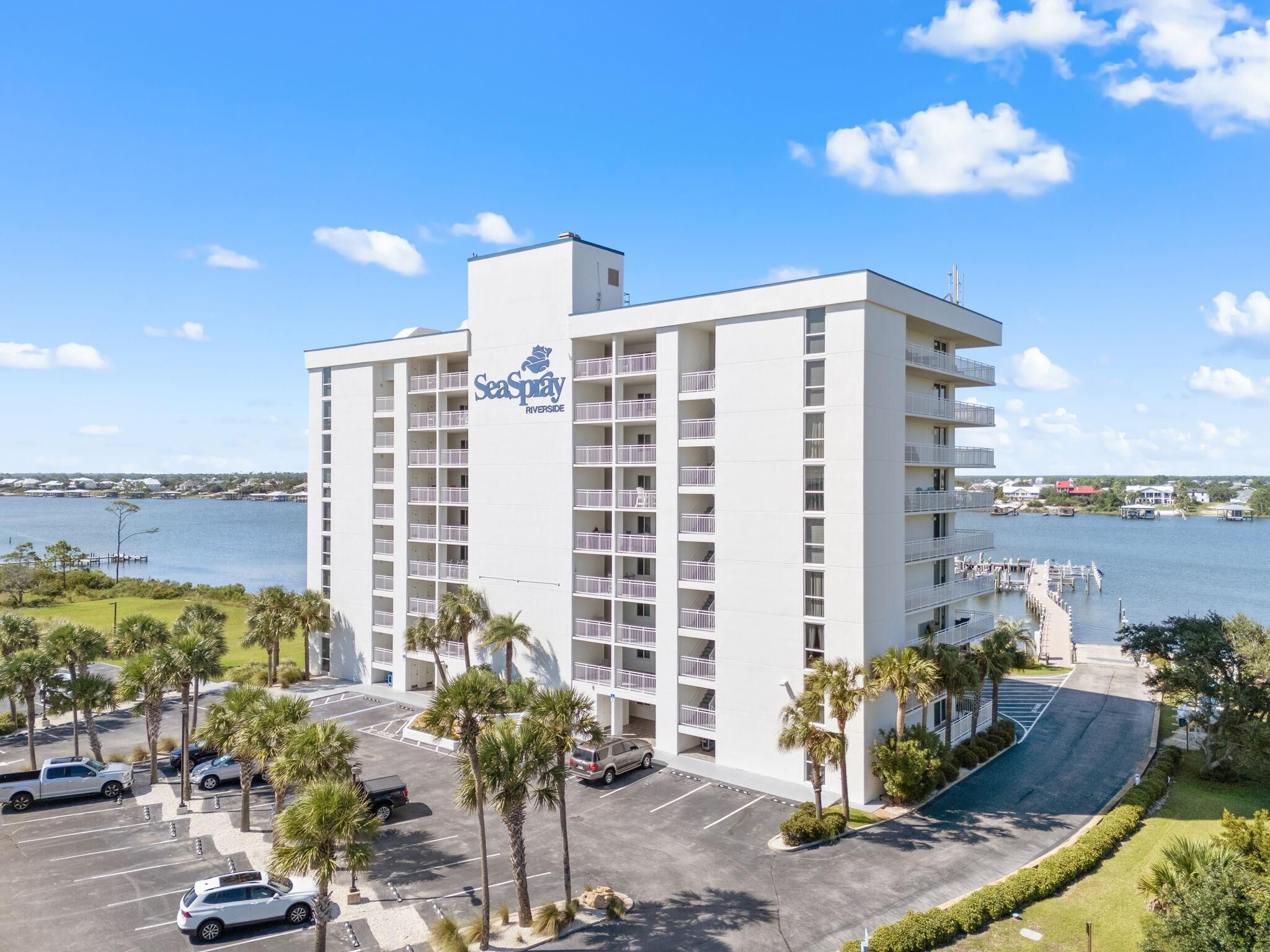 A multi-story hotel with balconies, a parking lot, and a view of the water.