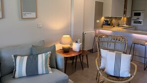 Una sala de estar moderna con un sofá gris, una silla de mimbre y una pequeña mesa de madera con una lámpara. Se ve una zona de cocina al fondo, con una encimera y electrodomésticos.