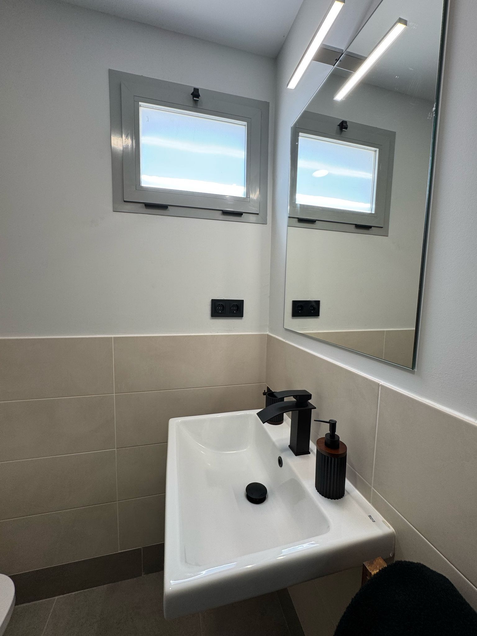 Un bagno moderno con lavandino bianco, rubinetto nero e specchio sopra. Due luci a parete e una finestra sopra lo specchio.