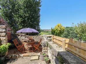 Eine Terrasse im Freien mit einer Steinmauer, einem Holztisch mit zwei Stühlen, einem Sonnenschirm und Blick auf die Landschaft.