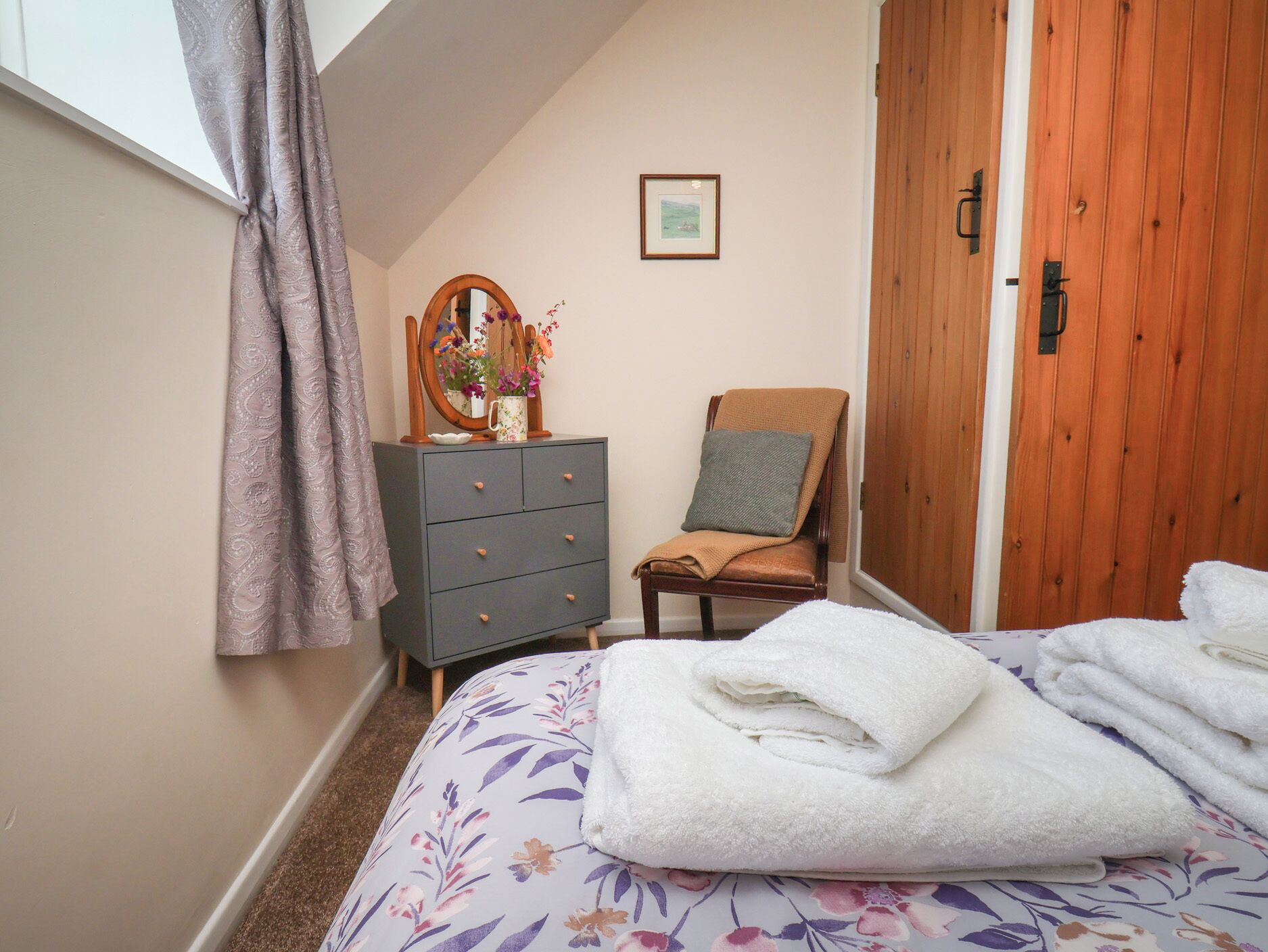 A bedroom with a bed, a dresser, a chair, a wooden door, and a window with curtains.