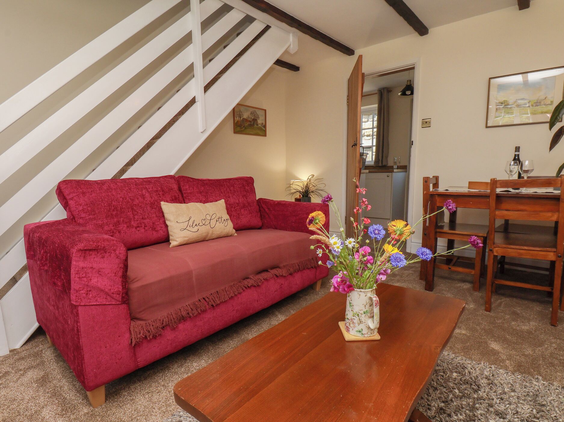 A red sofa with a cushion, a wooden coffee table with a floral vase, a wooden dining table with chairs, and a staircase with white railings.