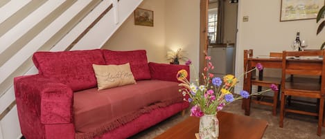 A red sofa with a cushion, a wooden coffee table with a floral vase, a wooden dining table with chairs, and a staircase with white railings.
