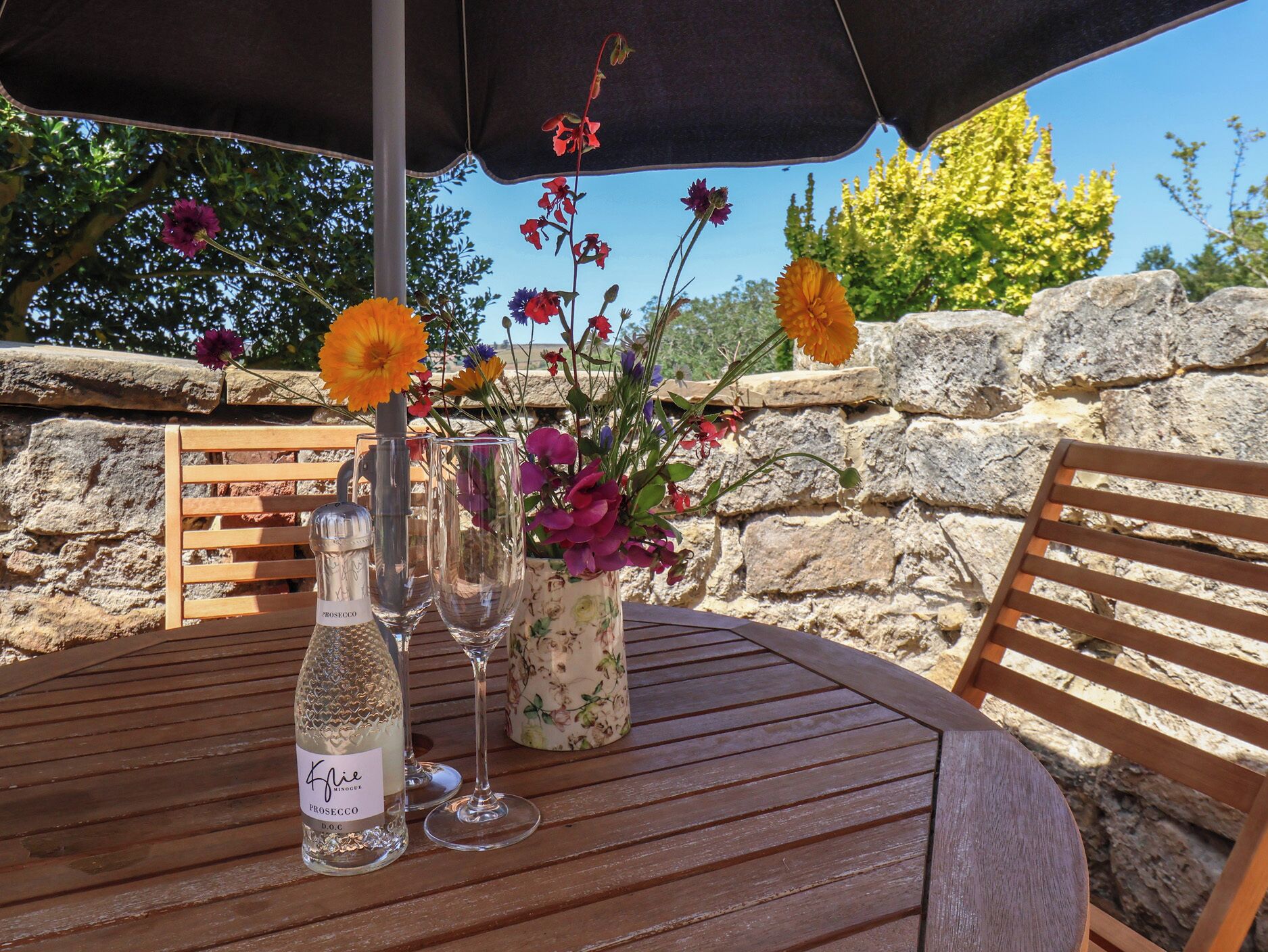 A wooden table with a bottle of Prosecco, two wine glasses, and a floral arrangement.