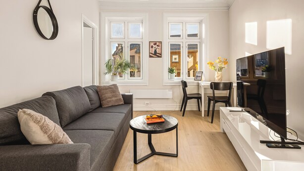 Een moderne woonkamer met een grijze bank, een klein rond salontafeltje, een televisie en een eetgedeelte met een witte tafel en stoelen.