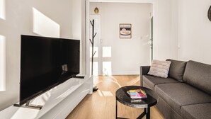Een moderne woonkamer met een flatscreen televisie, een grijze bank, een kleine ronde tafel met een boek, en een deur die naar een andere kamer leidt.
