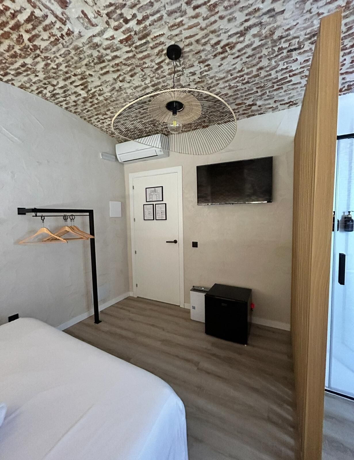 Ett hotellrum med en säng, ett klädställ, en takfläkt, en tv som är monterad på väggen och ett litet svart skåp.