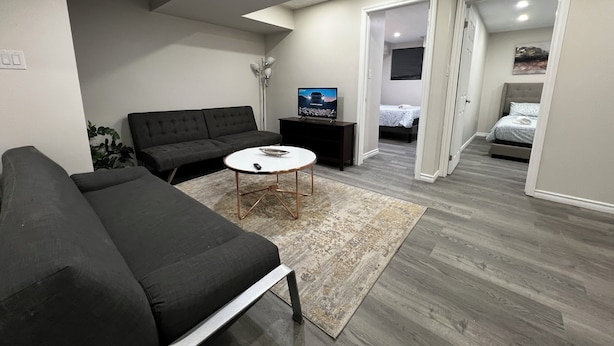 Een moderne woonkamer met een bank, een salontafel en een televisie. Er is een gang die leidt naar een slaapkamer met een bed en een deur.