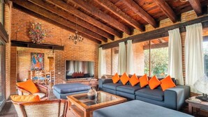 A rustic living room with exposed wooden beams, a brick fireplace, and a large sofa with orange cushions. - Villa Tinto : Private villa with pool and garden for relaxation and conviviality. (Cuernavaca)