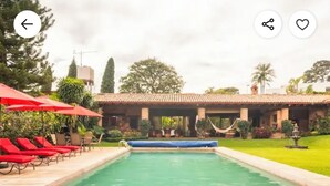 A swimming pool with red lounge chairs and umbrellas, surrounded by greenery and a building with a tiled roof. - Villa Tinto : Private villa with pool and garden for relaxation and conviviality. (Cuernavaca)