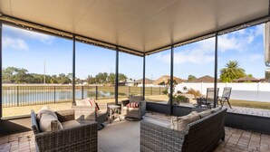 Un área de estar al aire libre techada con mobiliario de mimbre, una mesa y sillas, con vista a un cuerpo de agua y edificios residenciales.