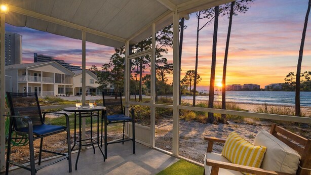 A covered outdoor seating area with a view of the sunset over the ocean, a building in the background, and a table with chairs. - Summer Lake 15- Shore to Please (Miramar Beach)