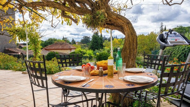 A round table set outdoors with plates, cutlery, and bottles, surrounded by chairs and a tree.