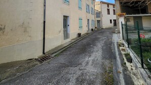 A narrow street with buildings on either side, a drainage grate in the middle, and a fence with plants on the right.