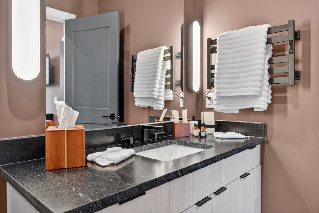 Une salle de bain moderne avec un plan de toilette noir, un lavabo blanc et une boîte à mouchoirs en bois. On y trouve deux porte-serviettes avec des serviettes blanches et un miroir au-dessus du lavabo.