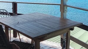 A covered outdoor seating area with a table and chairs, overlooking a calm sea and a distant hill. - New! Tikanot'la haut Exceptional view of Grande Anse beach (Deshaies)