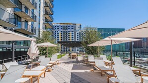 A rooftop lounge area with sun loungers and umbrellas, a small table, and a view of a modern building in the background. - Saiya  920 (Phoenix)