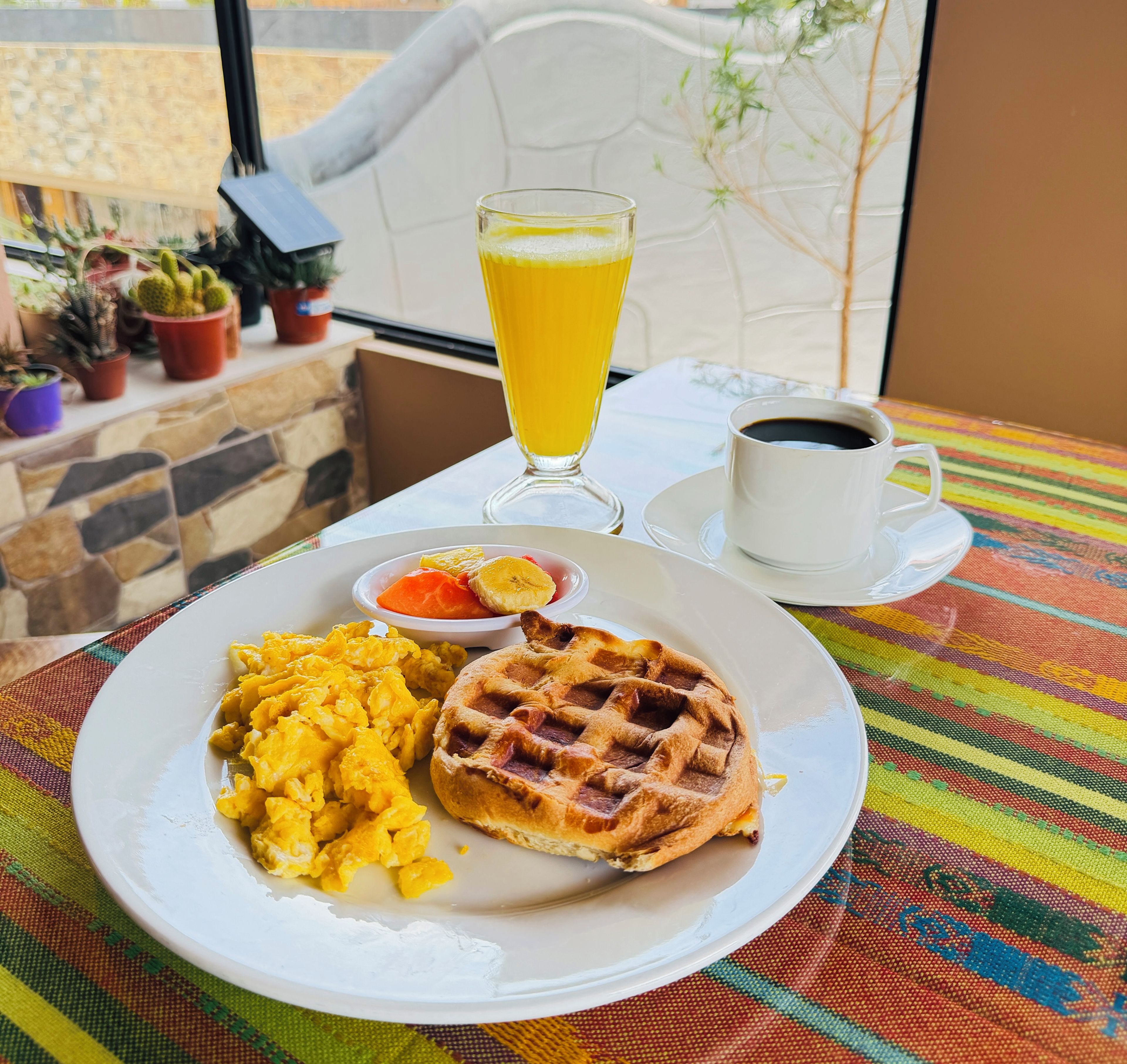 Un plato de desayuno con huevos revueltos, un waffle y un vaso de jugo de naranja sobre una mesa con mantel colorido.