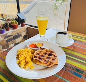 Ein Teller mit Rührei, einem Waffel und einem Glas Orangensaft auf einem Tisch mit farbenfroher Tischdecke.