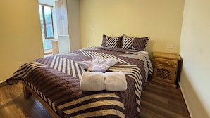 Een netjes opgemaakt bed met gestreept beddengoed en een opgevouwen handdoek, een houten nachtkastje met een lade, en een raam met uitzicht op buiten.
