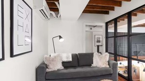 Ett modernt vardagsrum med en grå soffa, vita, fluffiga kuddar, en svart golvlampa och ett stort fönster med utsikt över omgivningen.
