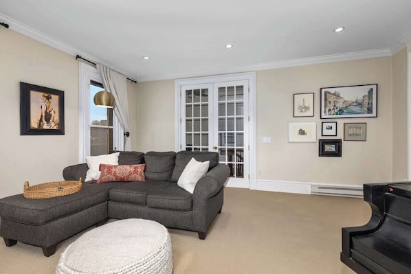 A living room with a sectional sofa, a round ottoman, a piano, and framed artwork on the walls.