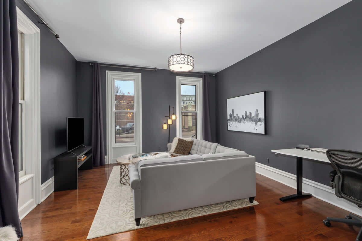 A modern bedroom with a grey sofa, a small table, a chair, and a cityscape artwork on the wall.