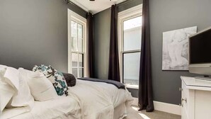 Une chambre à coucher avec un lit, une commode, un téléviseur et deux fenêtres avec des rideaux.