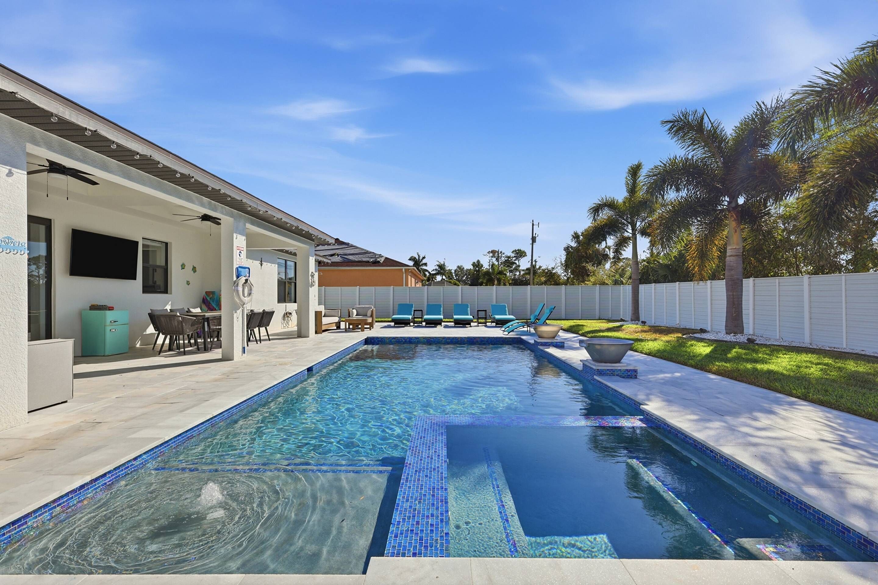 Une piscine avec un bord carrelé, un patio couvert équipé de chaises et d’une table, et une clôture blanche qui entoure la propriété.