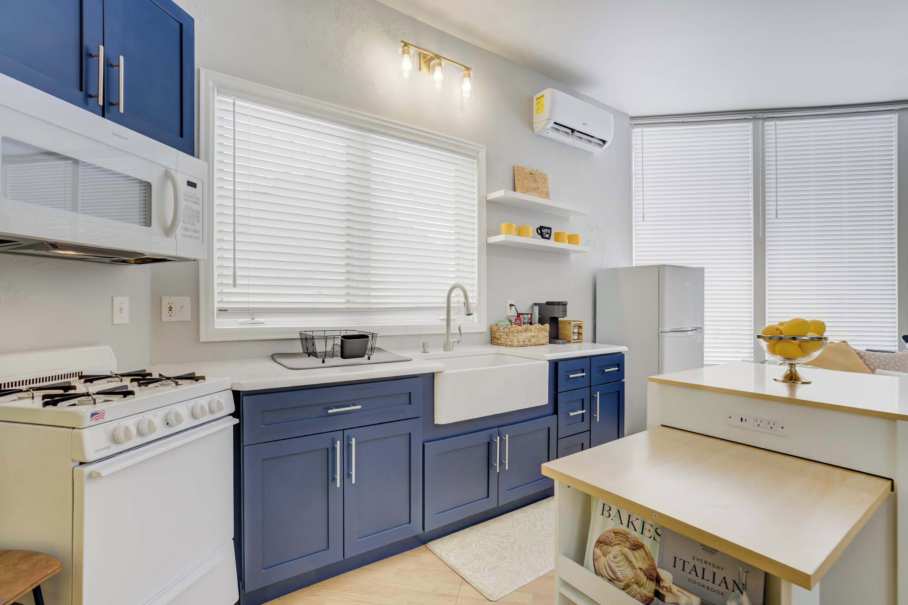 A modern kitchen with blue cabinets, a white stove, and a white countertop. There is a window with blinds, a sink, and a microwave above the stove.
