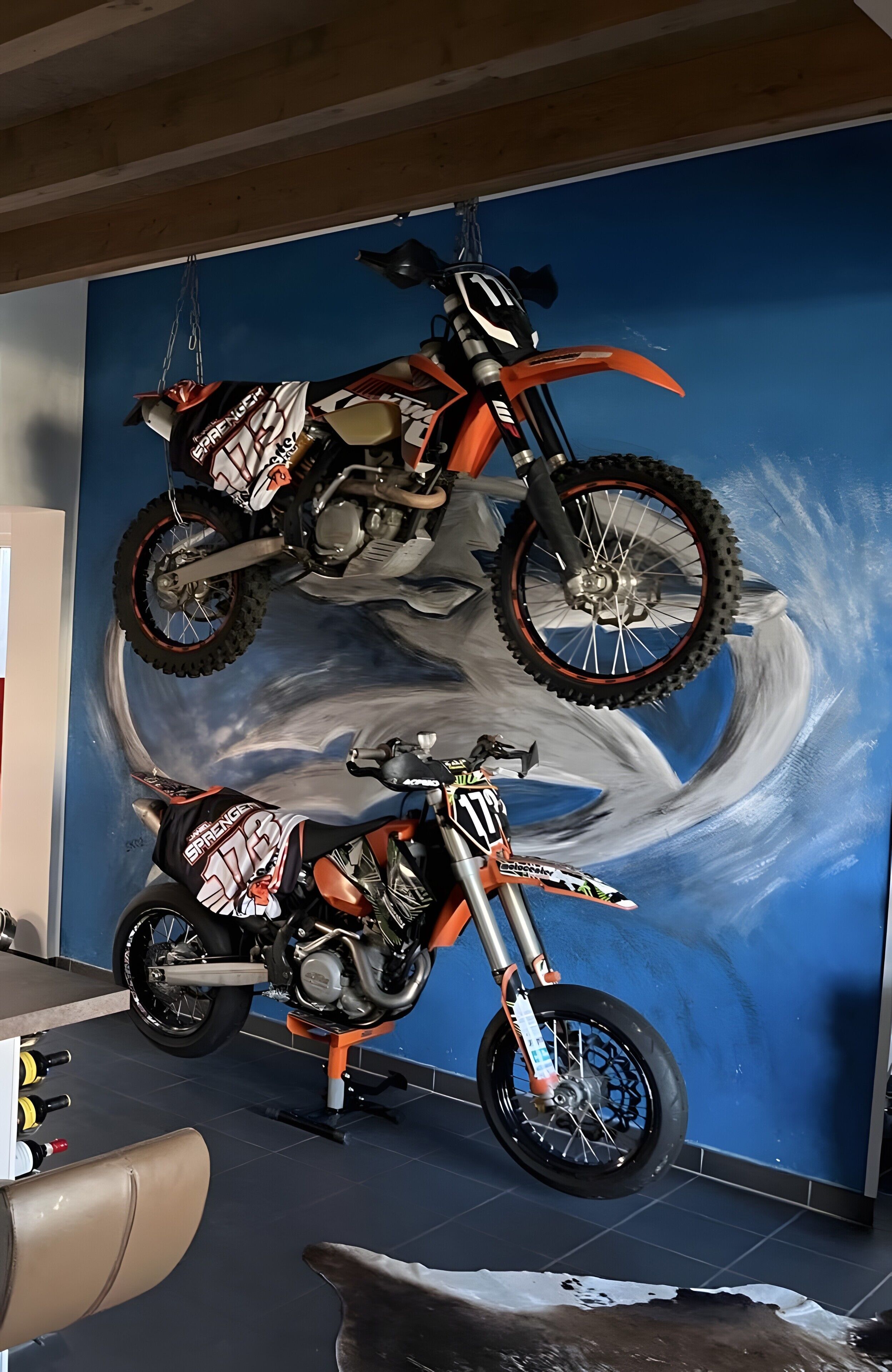 A motorcycle on a lift, a mural with a blue sky and clouds, and a chair in the foreground.