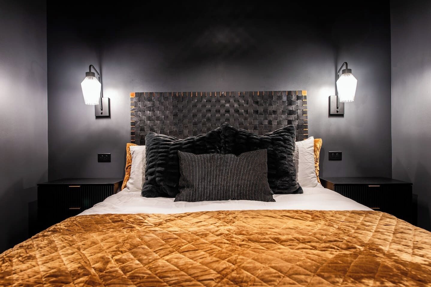 A modern bedroom with a quilted bedspread, a textured headboard, and two wall-mounted light fixtures.