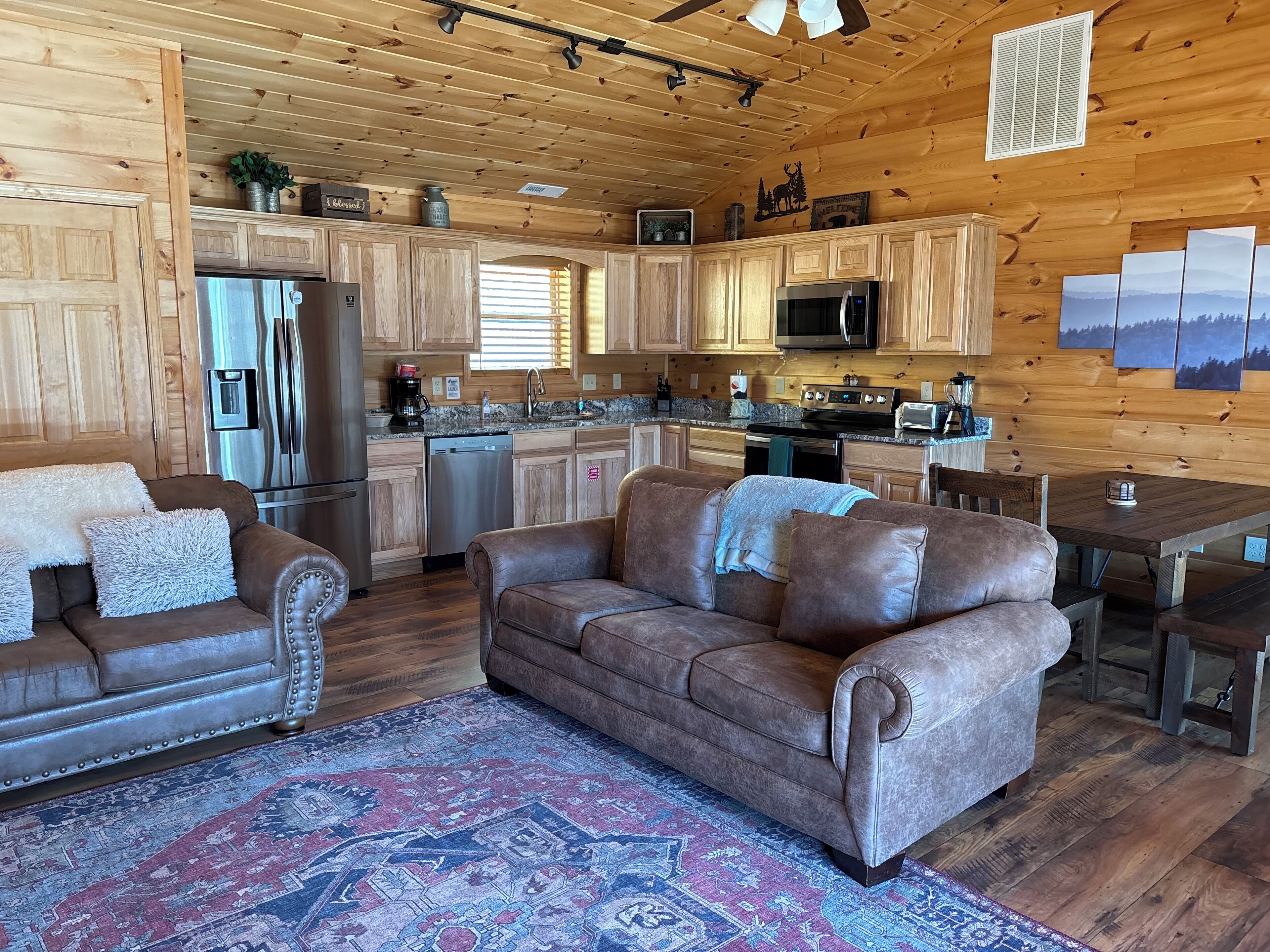 A cozy cabin interior with wooden walls, a dining area, a kitchen with modern appliances, and a large patterned rug.