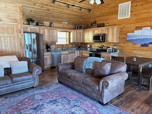A cozy cabin interior with wooden walls, a dining area, a kitchen with modern appliances, and a large patterned rug. - May the Forest Be with you (Gatlinburg)