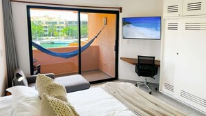 Habitación de hotel con cama, escritorio, televisión y vistas a una piscina y edificios a través de una puerta de vidrio.