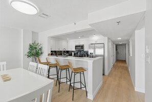 Eine moderne Küche mit weißen Schränken, einer zentralen Kochinsel mit Barhocker und einem Essbereich mit weißem Tisch und Stühlen.