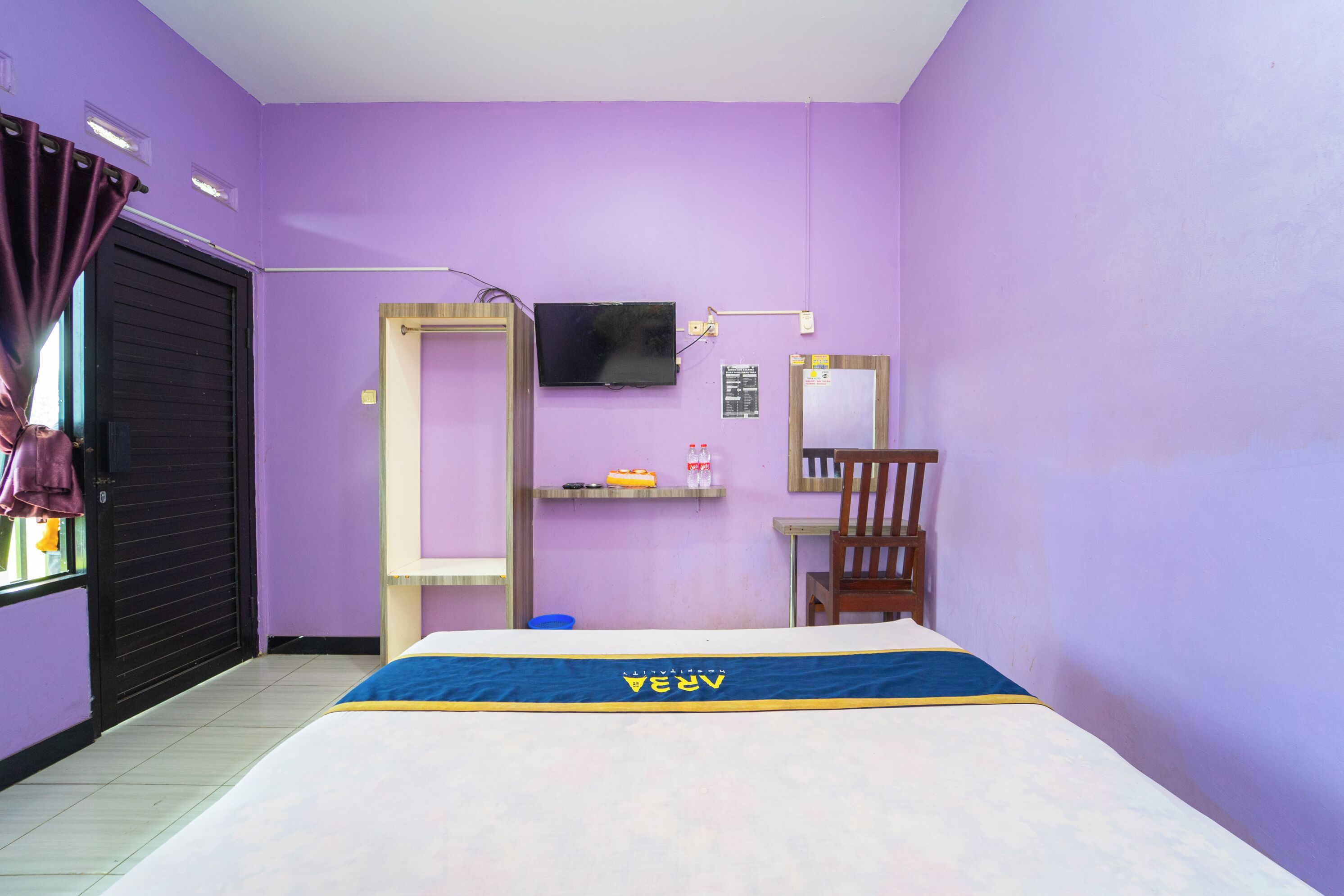 Una habitación con paredes moradas, una cama con colcha azul y amarilla, un televisor montado en la pared, una silla de madera y una pequeña estantería con objetos.