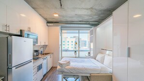 A compact hotel room with a kitchenette, a bed with a mattress and pillows, a small bedside table, and a view of the city through large windows. - Kenect 916 (Phoenix)