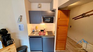 Uma cozinha compacta com micro-ondas, pia e um pequeno frigorífico. Há uma porta de madeira à direita e uma máquina de café à esquerda.