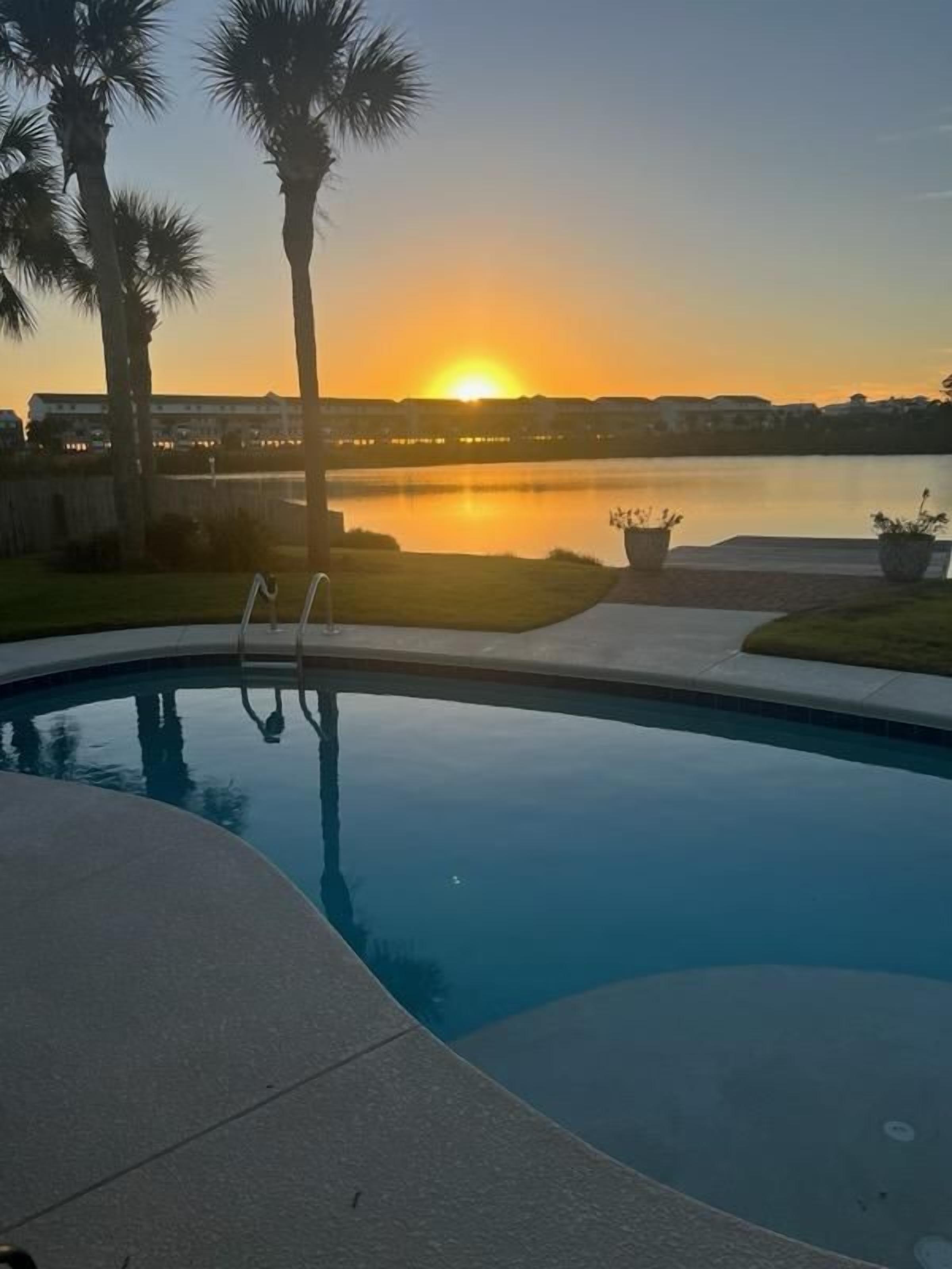 Une piscine offrant une vue sur le coucher de soleil au-dessus d’une étendue d’eau, entourée de palmiers et d’une zone herbeuse.