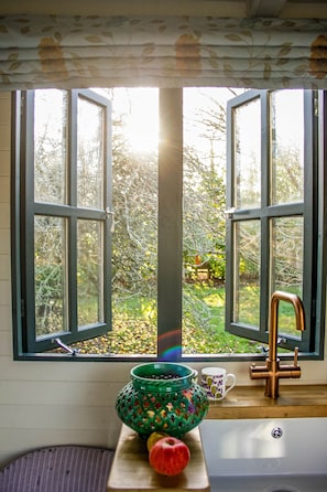 A window with a view of a garden, a green patterned bowl, a red apple, and a wooden chair. - Kent Family Retreat · Stable + Shepherd's Hut (Cranbrook)