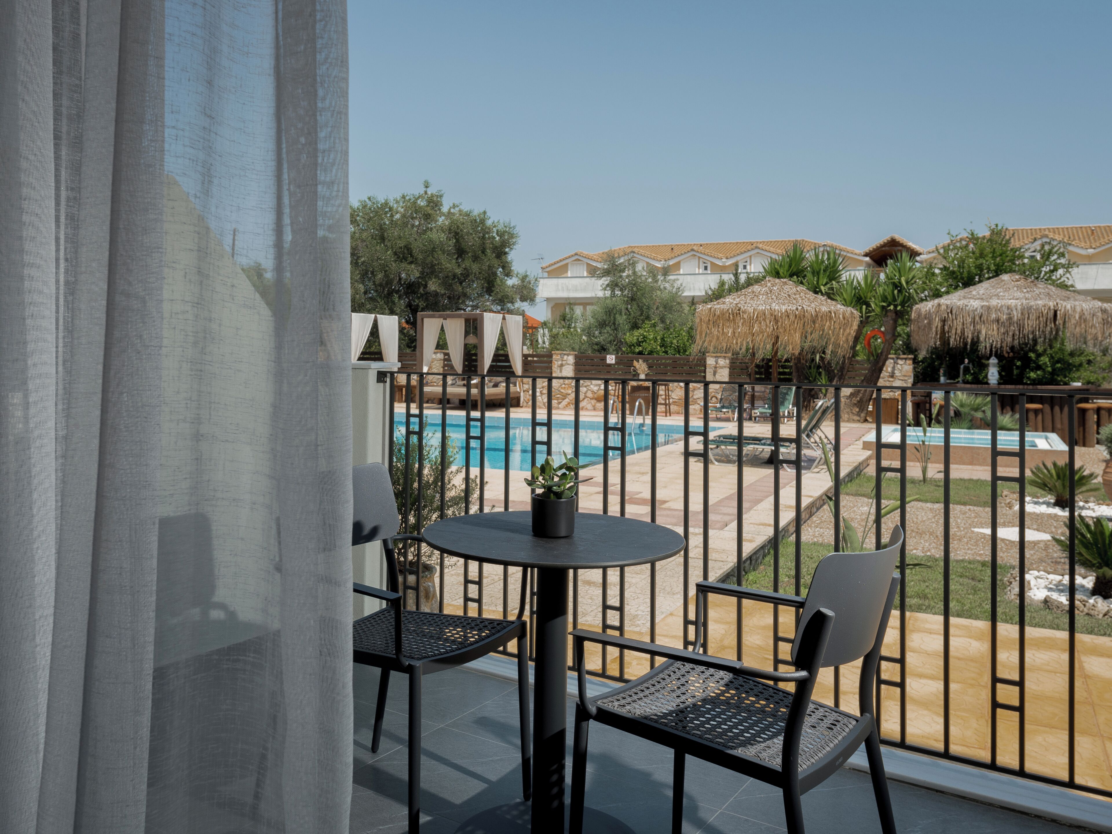 A balcony with a pool view, featuring a table and two chairs, a thatched umbrella, and a view of a pool and buildings.