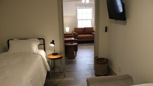 Hotellihuone, jossa on kaksi sänkyä, pieni pöytä lampun kanssa, sohva ja seinälle asennettu televisio.