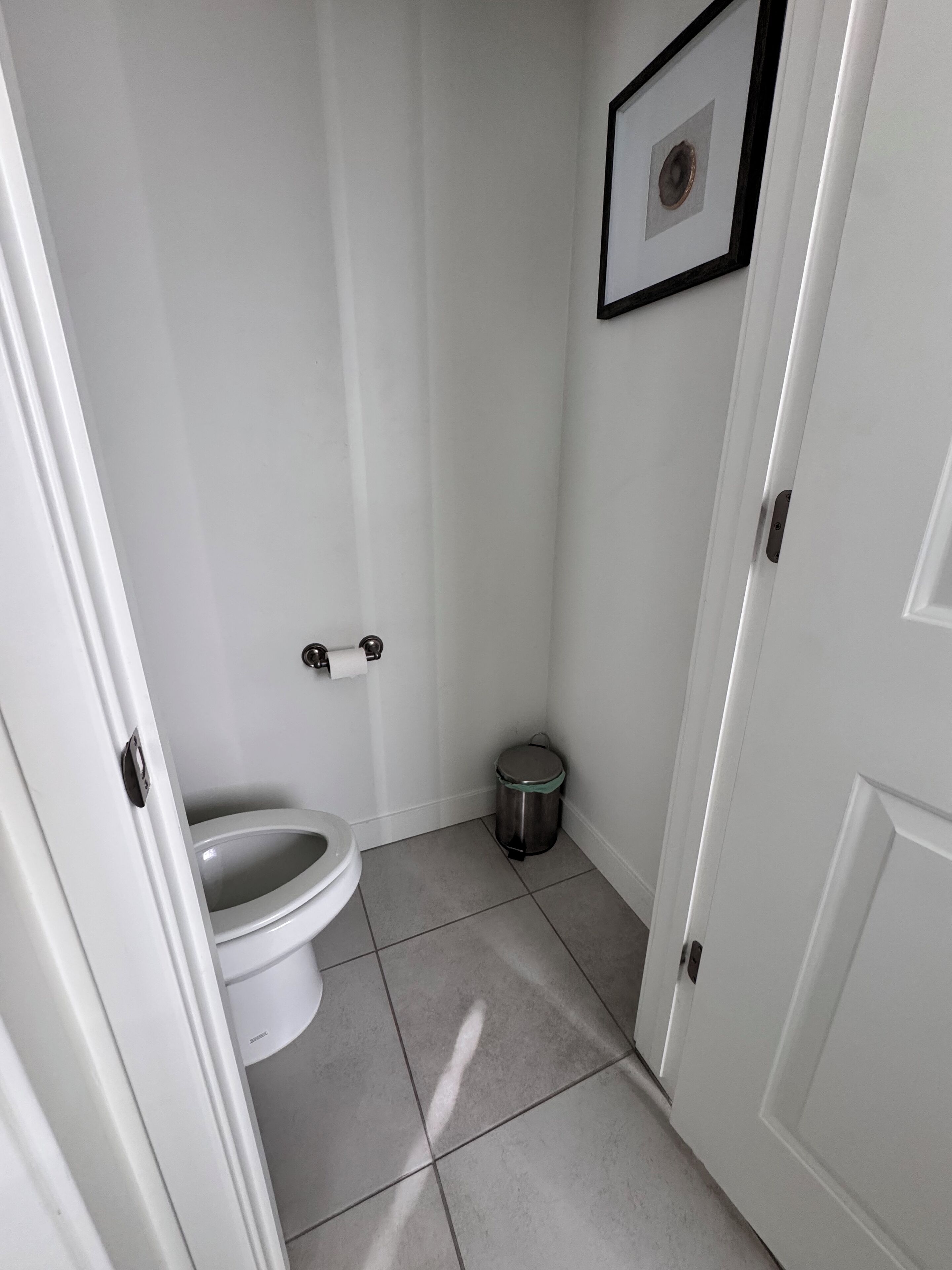 Un baño pequeño y limpio, con inodoro, cubo de basura y un cuadro en la pared.