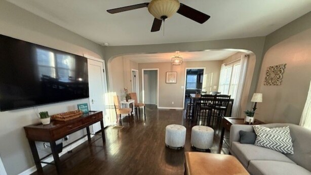 A spacious living room with a large flat-screen TV, a ceiling fan, and a dining area in the background.