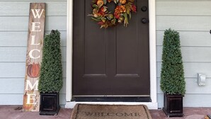 A front door with a dark brown door, a welcome wreath, a 'WELCOME' sign, and two small potted plants on either side.