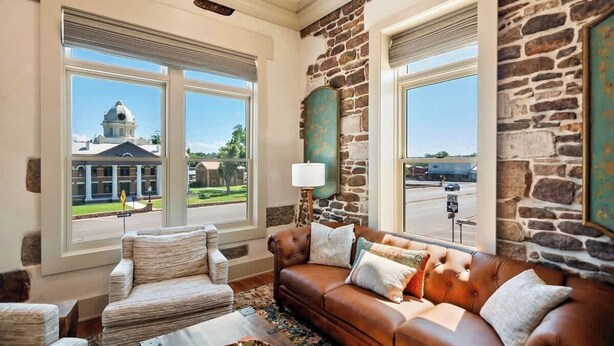 A living room with a brown leather sofa, a patterned armchair, and a coffee table. There are two windows with a view of a building and a street. - Drifter Suites Combo (Mason)