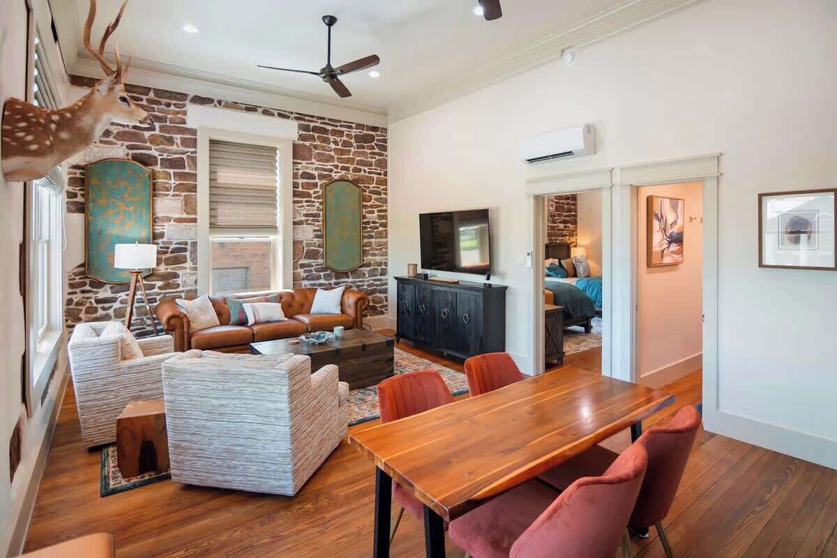 Un salon avec un mur en pierre, une tête de cerf accrochée au mur, un ventilateur de plafond, un téléviseur et un coin repas avec une table et des chaises en bois.