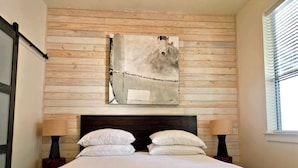 Une chambre avec un mur recouvert de lambris en bois, une grande peinture abstraite, une tête de lit sombre, deux tables de chevet avec des lampes et une fenêtre avec des stores.